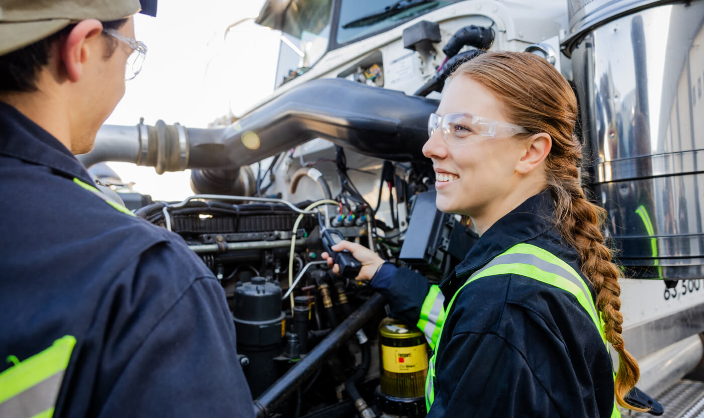 Female trades student with safety glasses smiling next to a hydrogen vehicle and a peer.