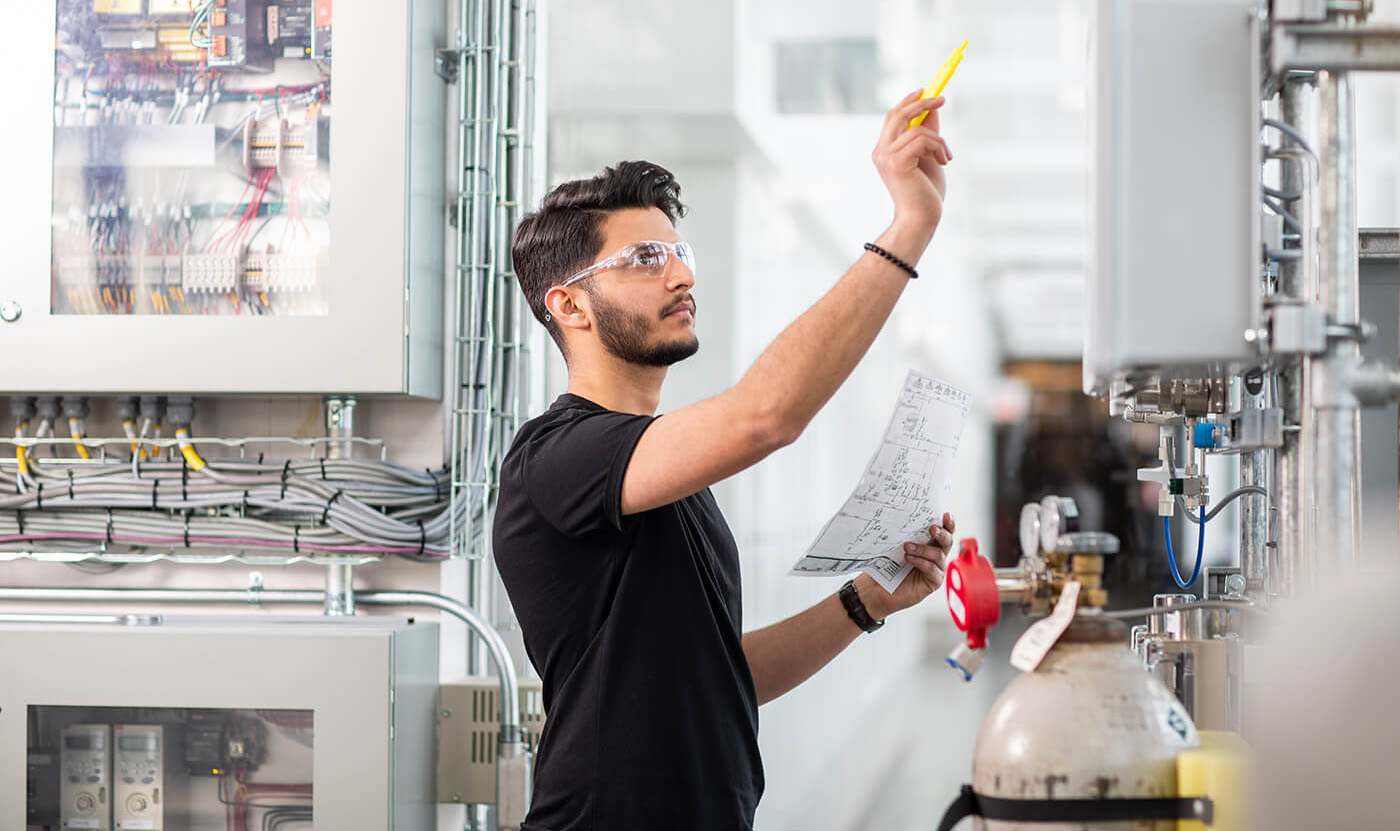 Male student in black shirt wearing safety goggles holds yellowhighlighter in air with right hand while doing a calculation