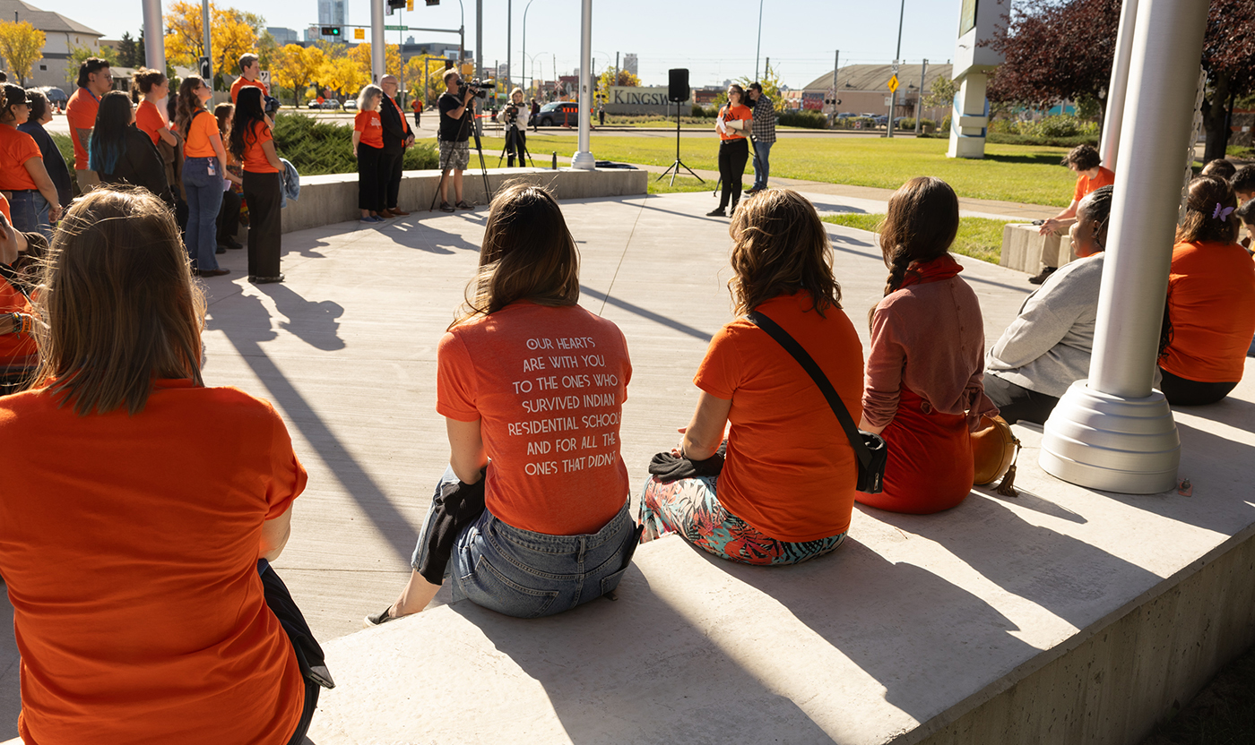 4 people sit outside wearing orange shirts watching flag lowering