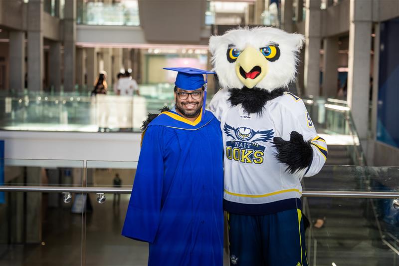 NAIT grad Justin Nand wears a convocation gown and cap with NAIT's Ook mascot