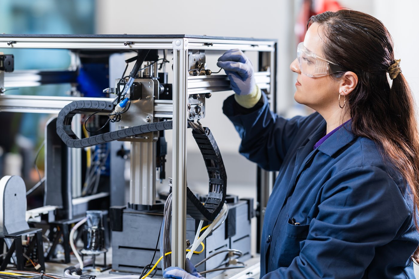 A person with long brown hair and protective eyeglasses looks into a piece of machinery