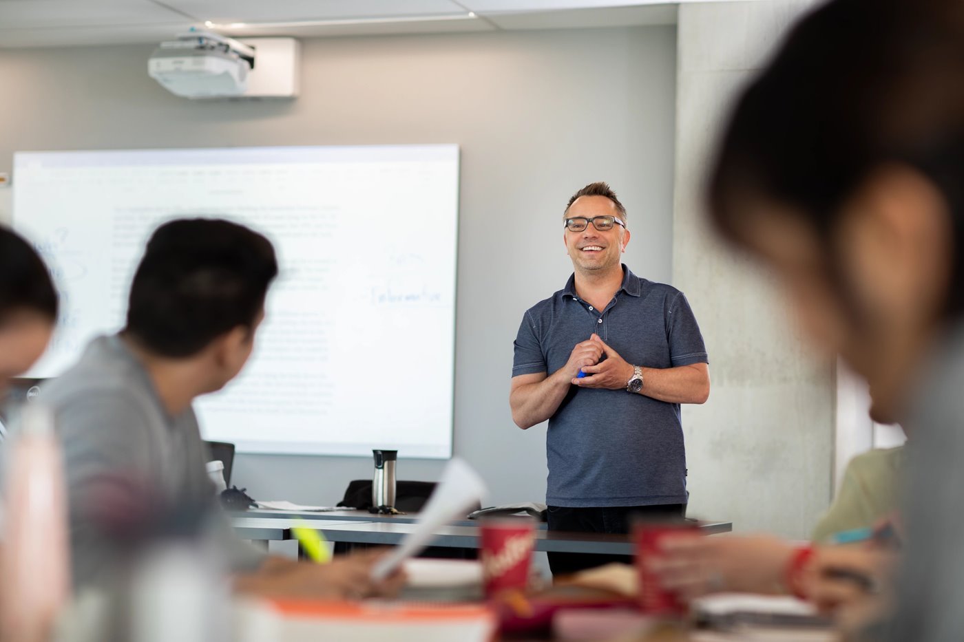 Male instructor standing at the front of a classroom, holding a whiteboard marker and smiling at his students