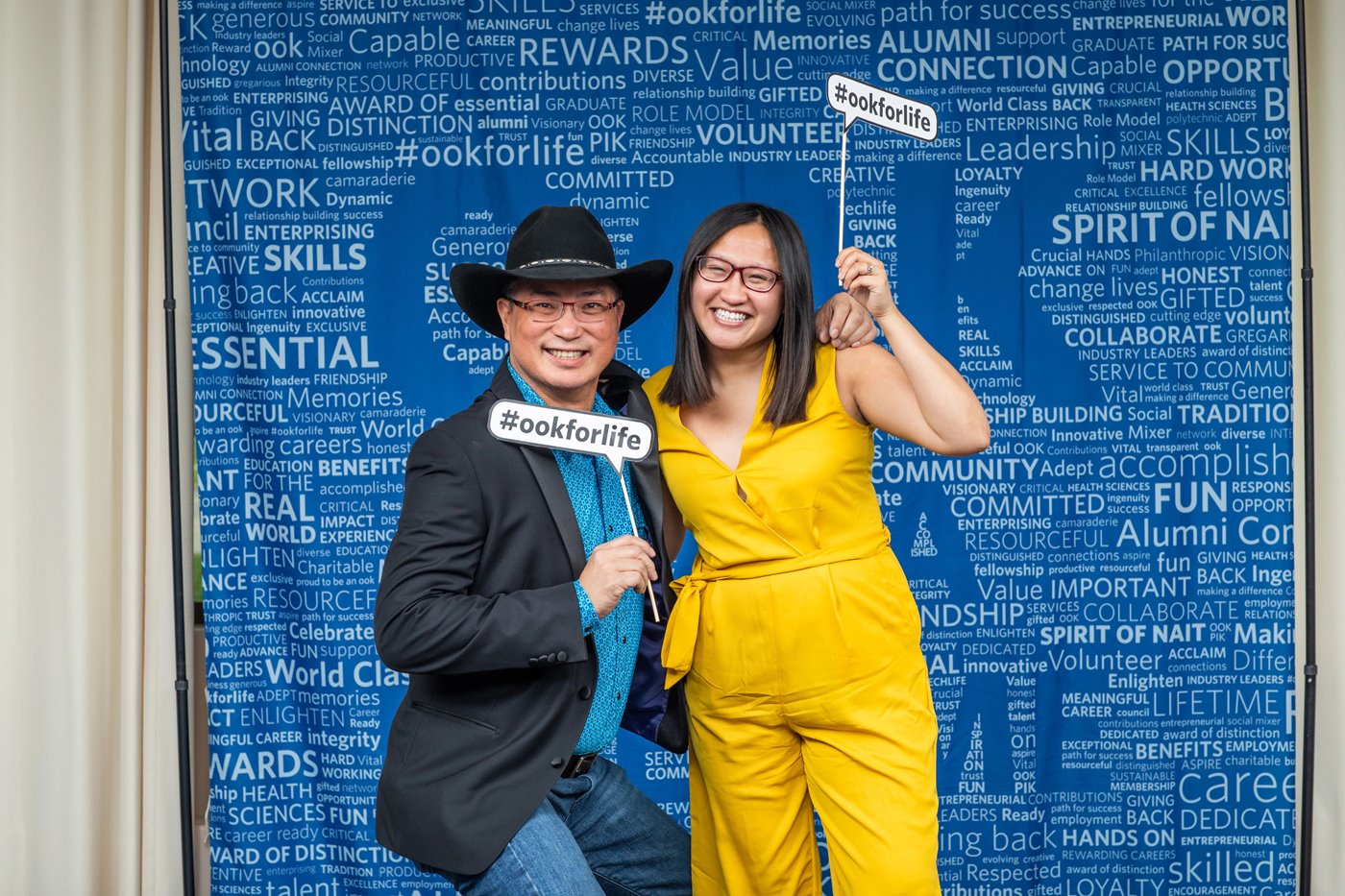 Linda Huang and Hubert Lau smile and pose at an alumni event in front of a blue NAIT alumni backdrop