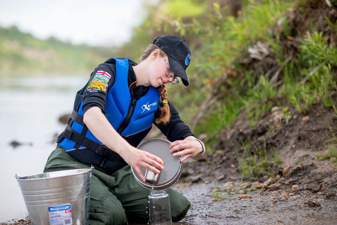 Woman kneeling on a river bank pouring water into a glass jar
