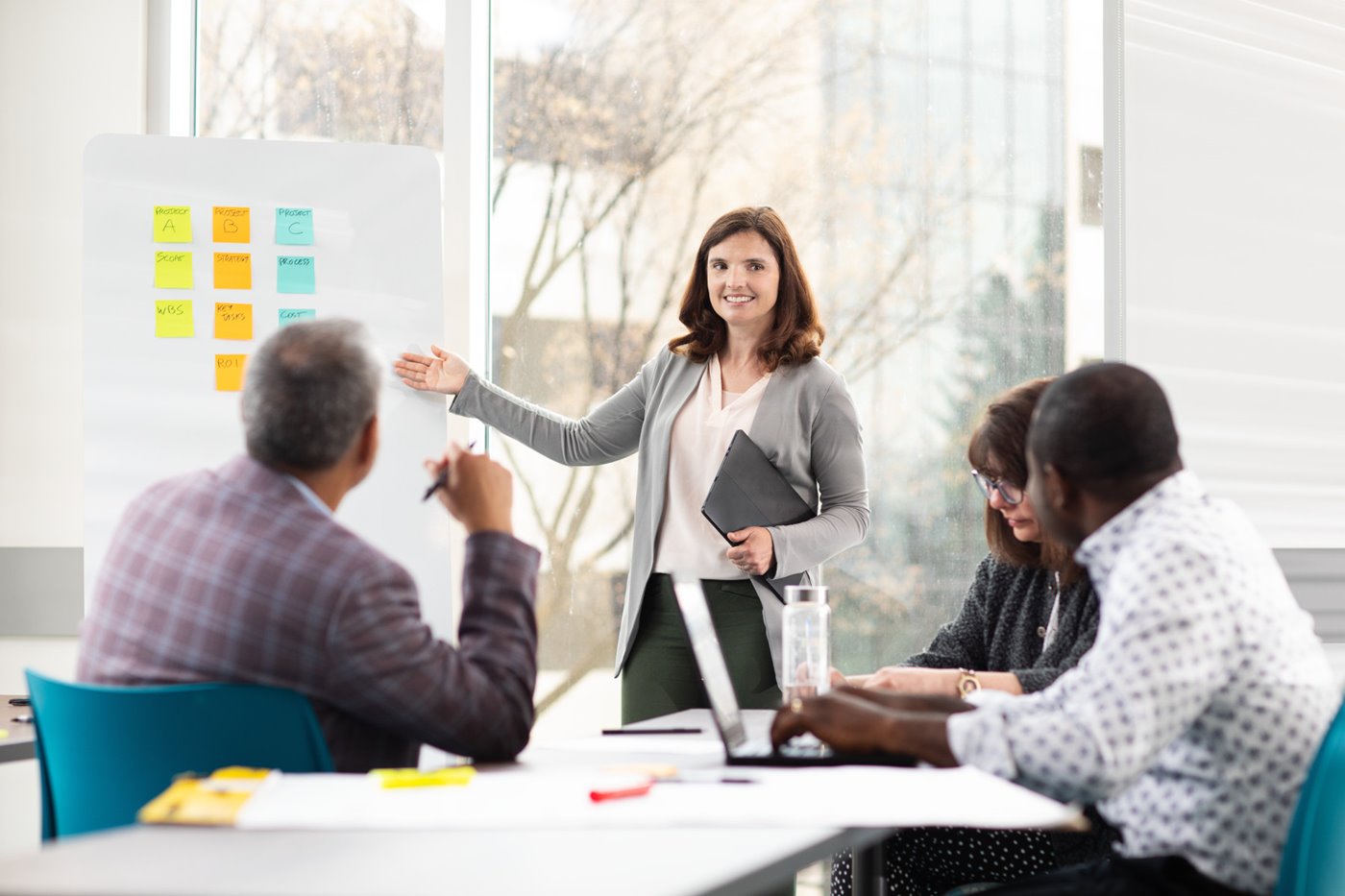 A woman gestures to a board with post-it notes while others sit and watch