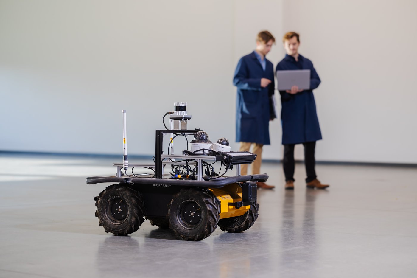 two individuals in blue lab coats look upon an autonomous drone with large treaded tires