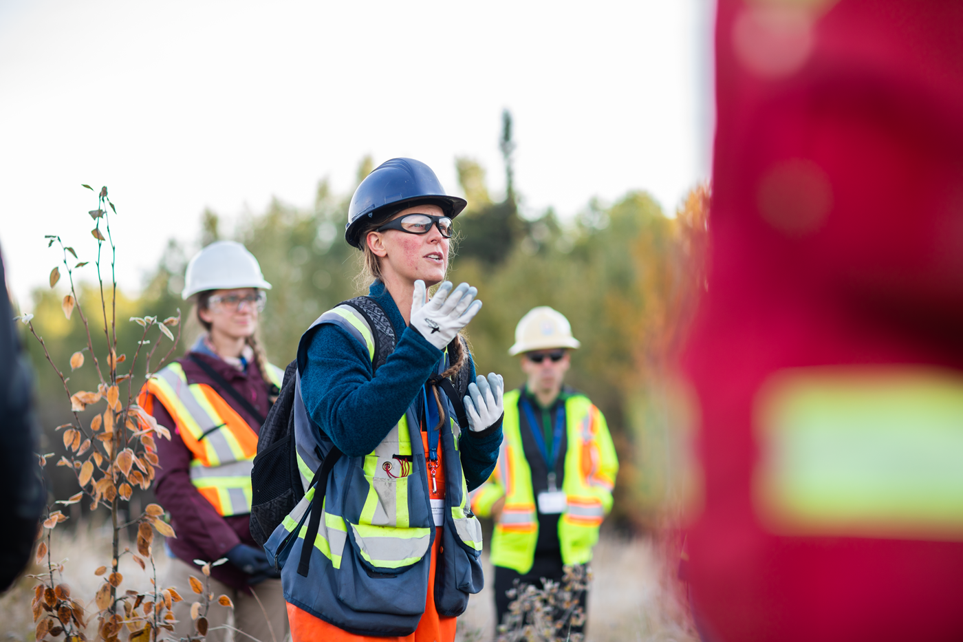 A person in safety gear is speaking to a crowd of people in a field within the boreal forest